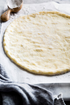 A rolled-out pizza dough on a floured surface with a wooden scoop and a kitchen towel nearby.