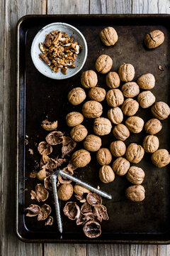 An overhead view of whole walnuts, cracked walnut shells, and a bowl of walnut pieces on a baking tray with a nutcracker.