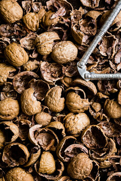 An assortment of whole and cracked walnut shells with a metal nutcracker on a dark surface.
