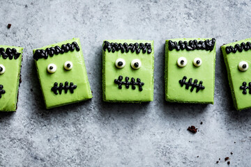 Five green Halloween-themed brownie squares decorated to resemble Frankenstein's monster, with candy eyes and black icing features, are arranged on a gray surface.