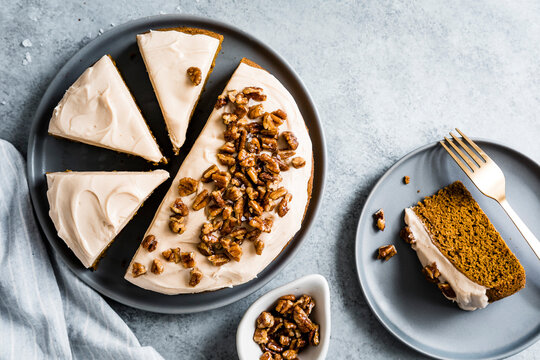 Top view of a sliced cake with frosting and pecans on a gray plate, alongside a slice on a separate plate with a fork.