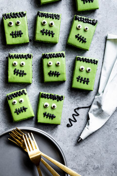 Green Frankenstein-themed cookies with black icing details are arranged on a gray surface next to a piping bag and gold forks on a plate.