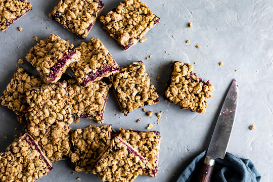 An assortment of berry crumble bars on a gray surface with a knife and cloth nearby.