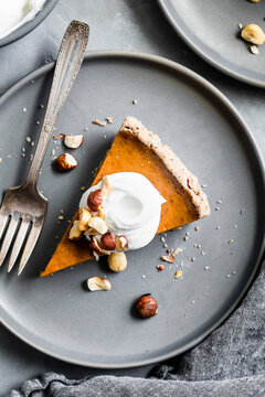 A slice of pumpkin pie topped with whipped cream and hazelnuts on a gray plate with a fork.