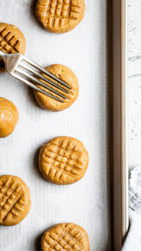Peanut butter cookies being prepared on a baking sheet with a fork creating a crisscross pattern.