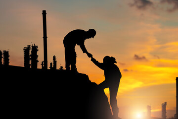 Silhouettes of Two workers helping each other up a hill with factory background, Engineer team people helping each other friend at Sunset Manufacturing Refinery Plant