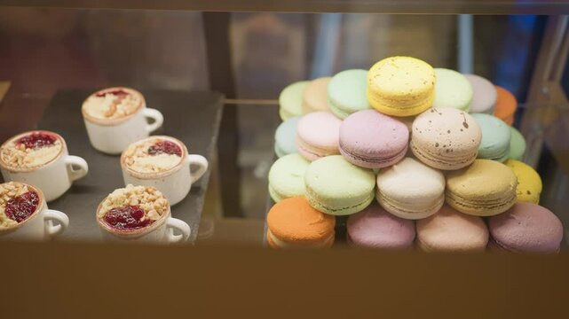 Close up of assorted colorful macarons arranged in pyramid shape beside small dessert cups with berry toppings and crumbles in store display case under warm lighting