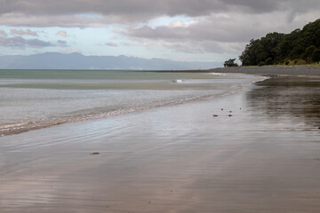 Calm waves wash ashore on a tranquil beach with trees and mountains under a cloudy sky, creating a peaceful coastal landscape. , Tapapakanga Regional Park, Auckland, New Zealand