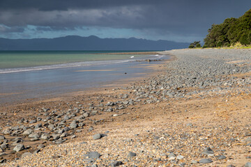 A rocky beach meets a calm sea under dark storm clouds approaching mountains along the New Zealand coast. Tapapakanga Regional Park, Auckland, New Zealand