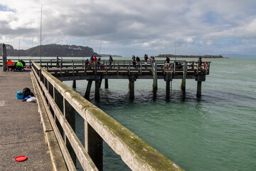 Obraz premium People of all ages gather on a pier in Waitawa Regional Park to fish, enjoying the coastal scenery and recreational activity. Kawakawa, Auckland, New Zealand