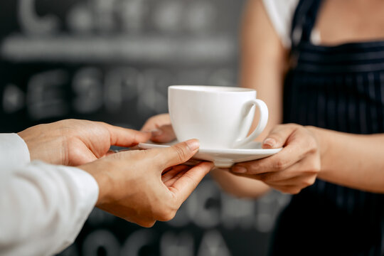 A young Asian female barista in a striped apron and orange shirt stands in a cozy modern café, smiling warmly as she hands a freshly made coffee cup to a woman customer. - Powered by Adobe