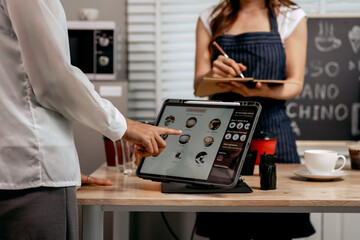 A young Asian female barista in a striped apron and orange shirt assists a woman customer choosing coffee from a tablet menu in a cozy modern café, offering espresso, cappuccino, latte, hot chocolate