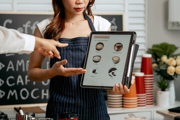 A young Asian female barista in a striped apron and orange shirt assists a woman customer choosing coffee from a tablet menu in a cozy modern café, offering espresso, cappuccino, latte, hot chocolate