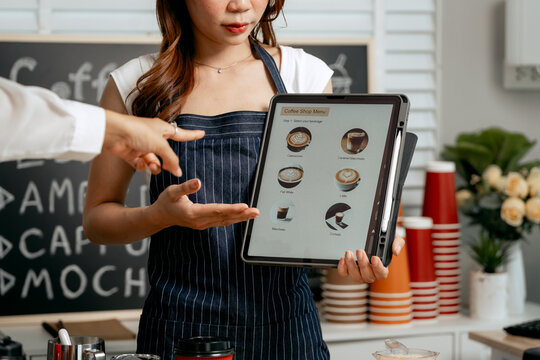 A young Asian female barista in a striped apron and orange shirt assists a woman customer choosing coffee from a tablet menu in a cozy modern café, offering espresso, cappuccino, latte, hot chocolate - Powered by Adobe