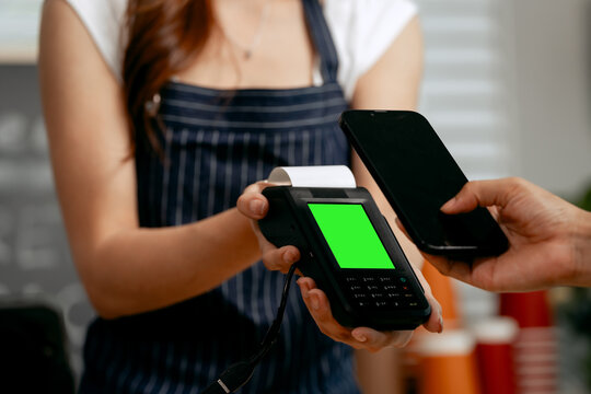 A young Asian female barista in a striped apron and orange shirt assists a woman customer making a quick, contactless payment via mobile QR code at the POS in a cozy modern cafe.
