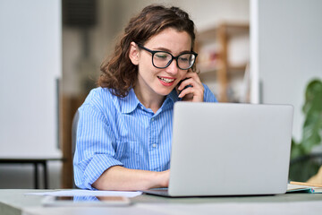 Busy young woman sales manager talking on cell phone using laptop working online on computer....