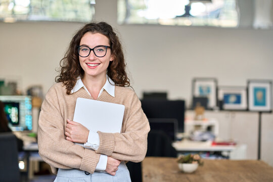 Happy businesswoman, female company worker intern or corporate manager holding laptop. Young smiling professional business woman standing in modern office working, looking at camera. Portrait