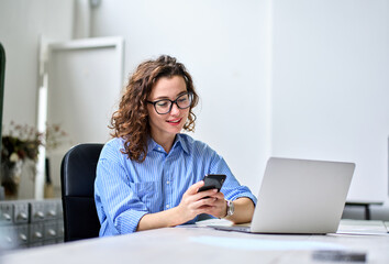 Busy young happy business woman, smiling professional businesswoman worker looking at smartphone using cellphone mobile technology apps working or in office checking cell phone sitting at desk.