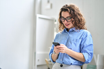 Smiling busy young business woman user, happy businesswoman wearing glasses holding cellular smartphone technology working standing in office using mobile phone working on cellphone looking at cell.
