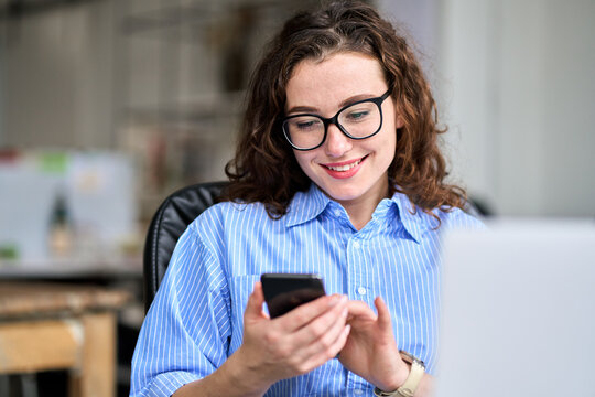 Happy professional female manager working in office typing on mobile cell phone sitting at desk. Young busy business woman looking at smartphone using cellphone mobile cell corporate tech device.