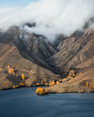 Autumn colored trees at the bottom of a mountain by a lake in the clouds.