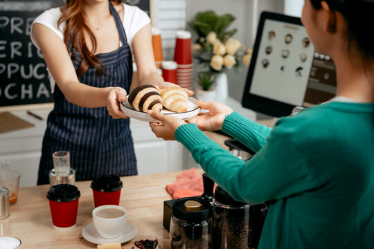A young Asian female barista in a striped apron and orange shirt warmly hands a freshly brewed coffee and a croissant to a woman customer in a cozy modern café, providing excellent service.