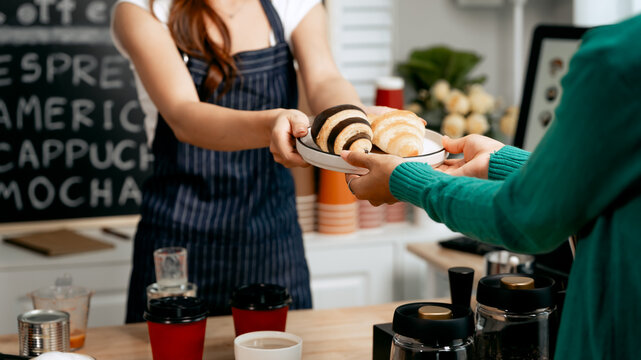 A young Asian female barista in a striped apron and orange shirt warmly hands a freshly brewed coffee and a croissant to a woman customer in a cozy modern café, providing excellent service. - Powered by Adobe