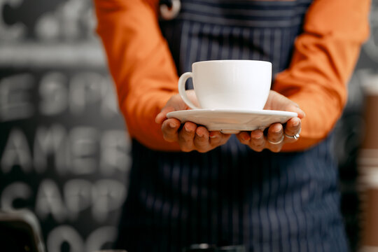 A young Asian female barista in a striped apron and orange shirt prepares a latte coffee for an online order in a cozy modern café, surrounded by takeaway cups and professional equipment.