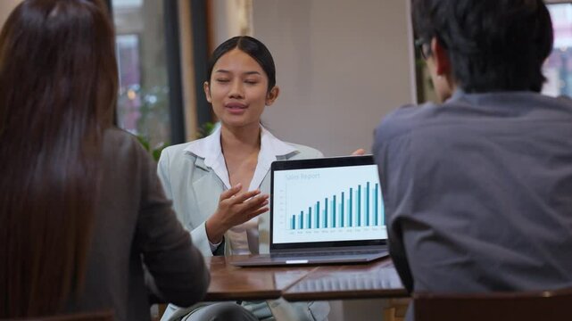 Young Asian businesswoman presenting a sales report to colleagues during a team meeting in a modern office. Confident female leader engaging in professional communication, explaining data and strategy - Powered by Adobe