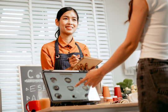A young Asian female barista in modern cafe assists woman customer ordering latte, fresh coffee beans, dark roast, croissant, completing the purchase with mobile payment at the POS using QR code. - Powered by Adobe