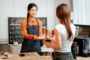 A young Asian female barista in modern cafe assists woman customer ordering latte, fresh coffee...