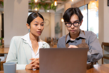 Young Asian businesswoman presents a marketing strategy on a laptop to a small business owner in a coffee shop. The cafe owner listens attentively while wearing an apron.