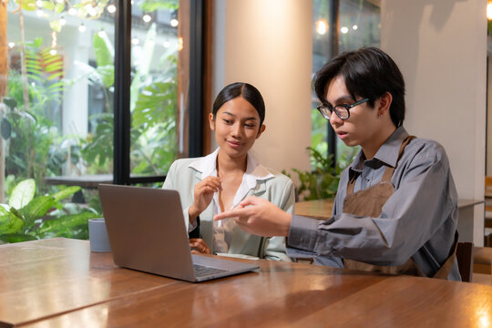 Young Asian businesswoman presents a marketing strategy on a laptop to a small business owner in a coffee shop. The cafe owner listens attentively while wearing an apron.