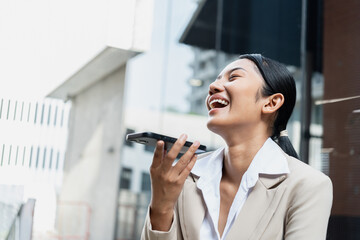 Asian businesswoman in a beige suit talking to voice assistant or chatbot on her smartphone outdoors, standing in modern city environment during daylight, using ai technology, smart assistant.