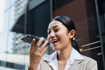 Asian businesswoman in a beige suit talking to voice assistant or chatbot on her smartphone outdoors, standing in modern city environment during daylight, using ai technology, smart assistant.