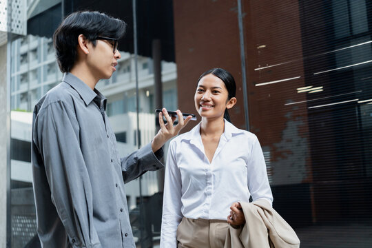 Young man holds a smartphone while a woman interacts with a voice-based AI chatbot assistant in an urban setting. Modern technology, artificial intelligence, and digital communication in daily life.