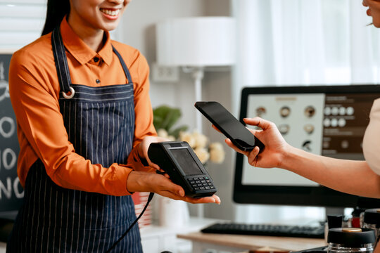A young Asian female barista in modern cafe assists woman customer ordering latte, fresh coffee beans, dark roast, croissant, completing the purchase with mobile payment at the POS using QR code.
