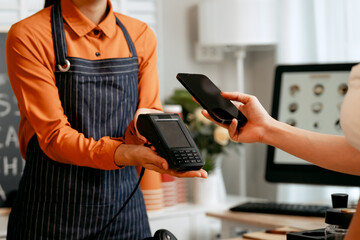 A young Asian female barista in modern cafe assists woman customer ordering latte, fresh coffee beans, dark roast, croissant, completing the purchase with mobile payment at the POS using QR code.