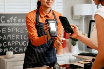 A young Asian female barista in modern cafe assists woman customer ordering latte, fresh coffee...