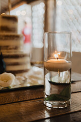 Floating candle centerpiece in glass vase with greenery on rustic wood table — romantic wedding decor with blurred cake in background, perfect for lifestyle, event, and celebration content