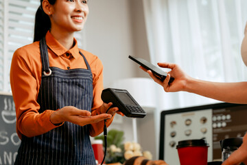 A young Asian female barista in modern cafe assists woman customer ordering latte, fresh coffee beans, dark roast, croissant, completing the purchase with mobile payment at the POS using QR code.