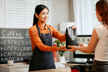A young Asian female barista in modern cafe assists woman customer ordering latte, fresh coffee...