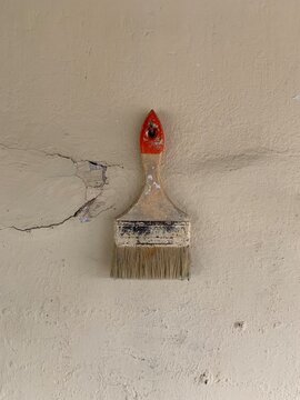 An old, worn paintbrush with frayed bristles leans against a damaged wall. The aged wall features cracks and a visible hole, evoking a rustic and weathered aesthetic.