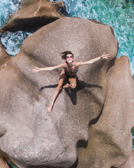 Aerial view of young energetic woman sitting on rocks formation with open arms towards camera and sky next to sea on La Dique island on Seychelles. Power of  traveling concept, drone
