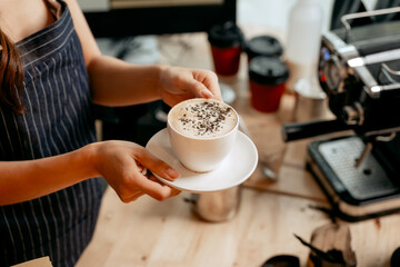A young female barista in a striped apron presents a freshly made cup of cappuccino topped with chocolate, holding it carefully on a saucer in a cozy, modern coffee shop setting.