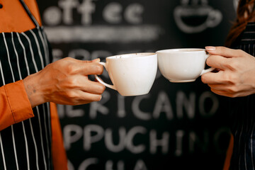Two young Asian female baristas in striped aprons and orange shirts enjoy teaching each other how...