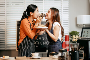 Two young Asian female baristas in striped aprons and orange shirts enjoy teaching each other how to make latte coffee, smell aroma, and taste coffee and croissants together in cozy modern café.