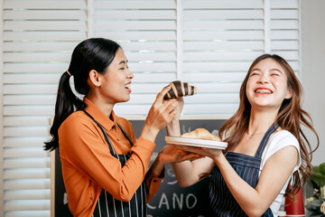 Two young Asian female baristas in striped aprons and orange shirts enjoy teaching each other how to make latte coffee, smell aroma, and taste coffee and croissants together in cozy modern café.