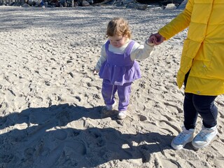 A child dressed in purple playfully walks on the sandy beach holding her mothers hand The sun...