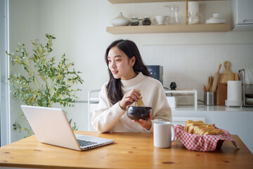 Young female preparing traditional matcha while using laptop in a bright modern kitchen.